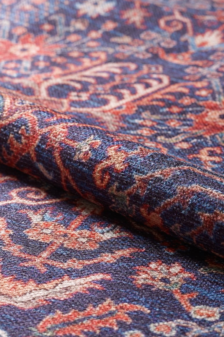 A close-up of the Solstice Red Navy Washable Rug (LCC3026), rolled up to showcase its intricate blue, red, and cream floral and geometric patterns.