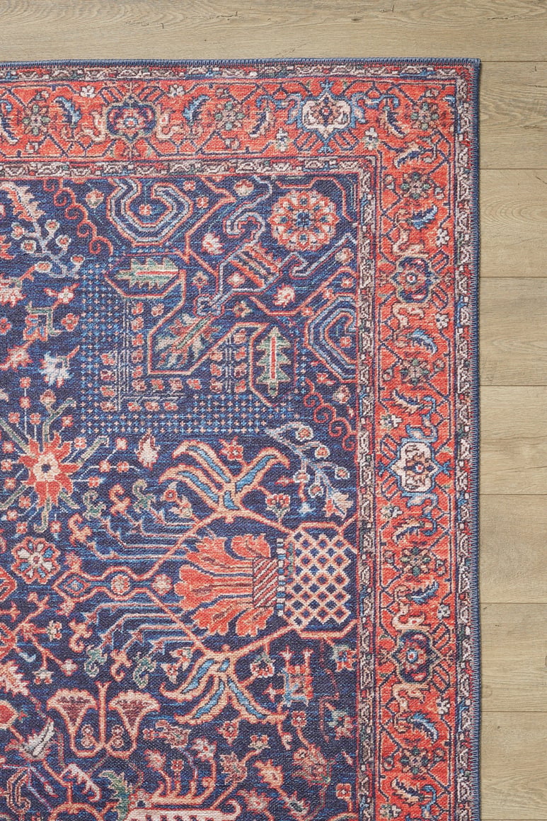 A close-up of the Solstice Red Navy Washable Rug - LCC3026 shows an intricate red, blue, and orange floral design, bordered by a wooden floor; this allergy-sensitive rug is easy to wash.