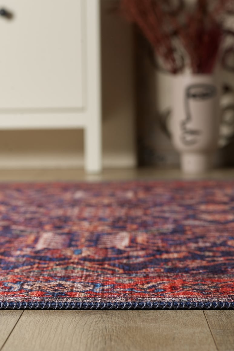Close-up of the Solstice Red Navy Washable Rug (LCC3026) featuring vibrant red, blue, and purple patterns on a wooden floor. A white cabinet and a decorative vase with dried plants enhance this allergy-friendly home accessory.