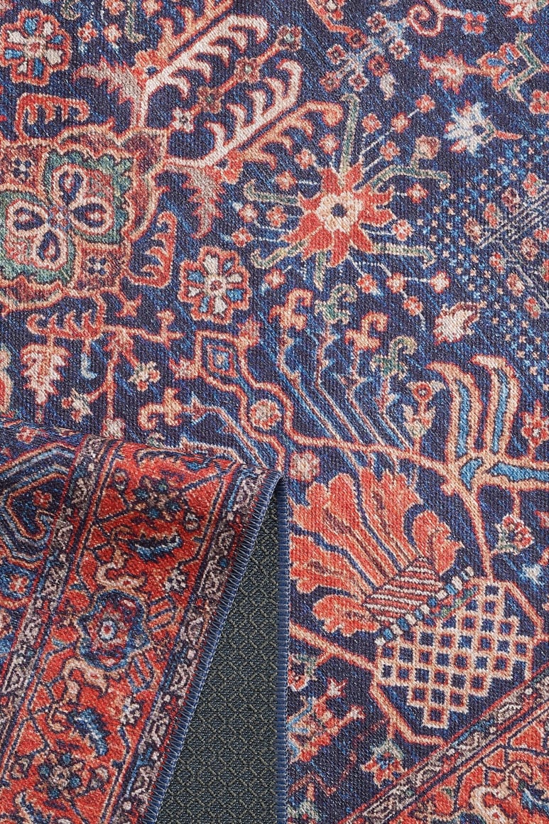 A close-up of the Solstice Red Navy Washable Rug - LCC3026 (Custom Size) shows blue with intricate red, orange, and cream floral and geometric designs. The folded corner reveals its textured underside—durable for high-traffic areas and easy to wash.