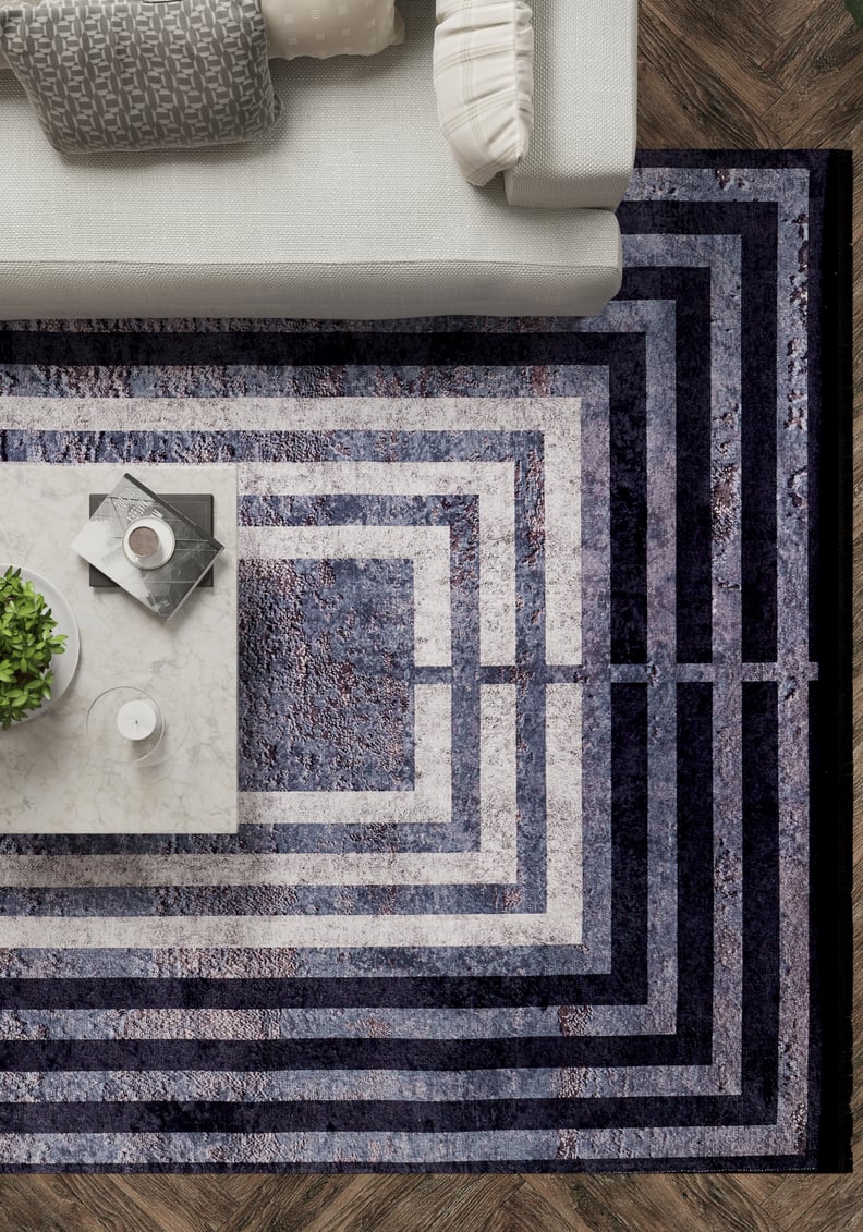 A modern living room corner features a light gray sofa, patterned cushions, a marble coffee table with a plant, book, and candle, all atop the Sophisticated and Chic - Washable Rug - JR1409 with geometric navy and white rectangles on a wooden floor.