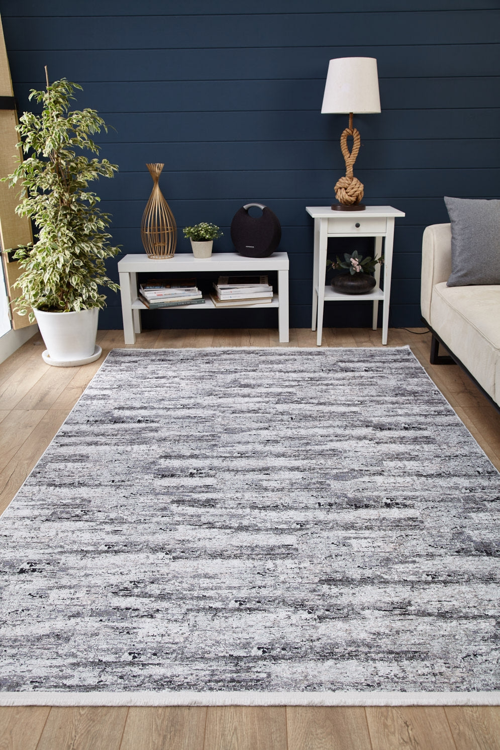 A modern living room showcases the Stone Path - Washable Rug - JR1772 (Custom Size) in gray-anthracite on wood floors, complemented by a beige sofa, white side table with lamp, plant, and navy blue accent wall—ideal for high-traffic spaces.