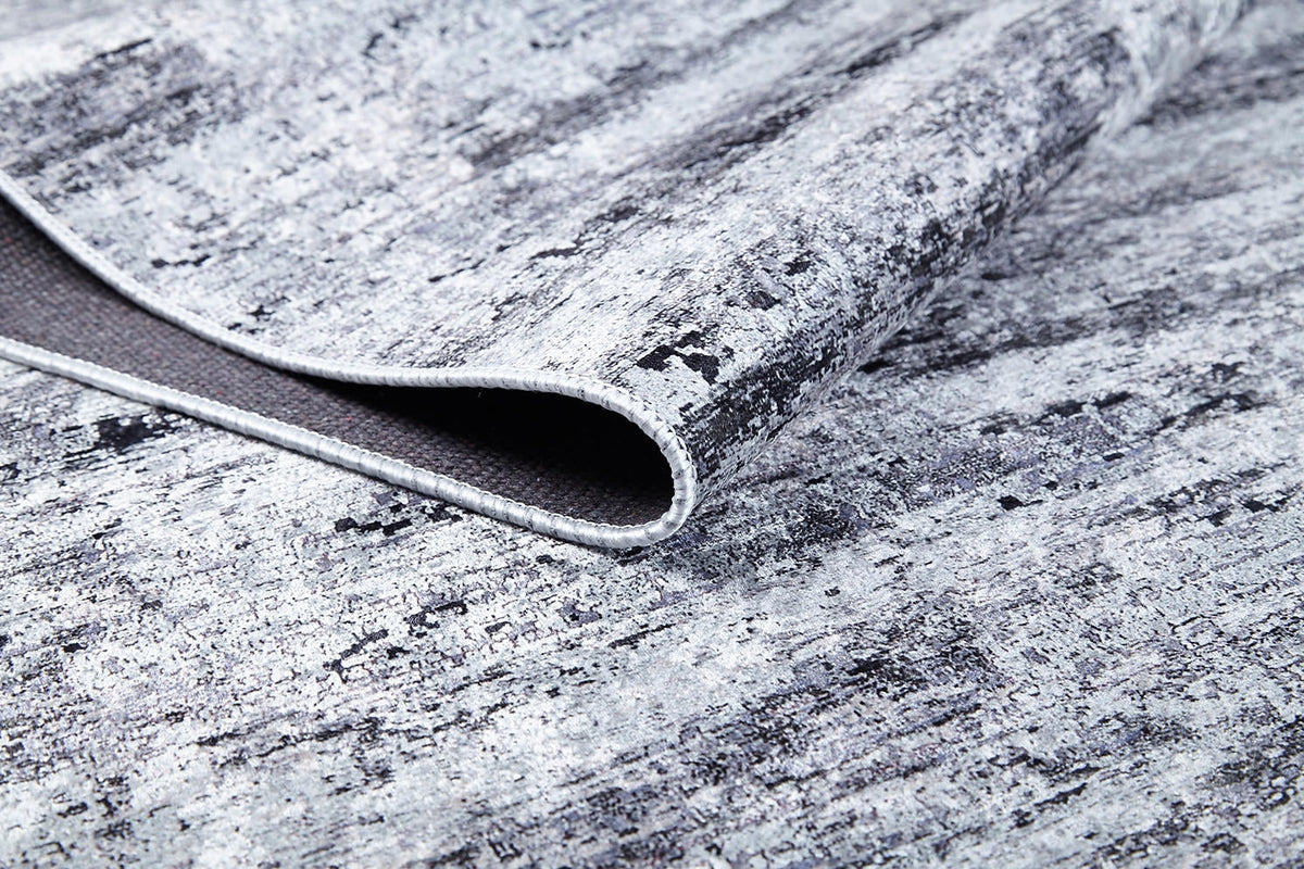 A close-up of the Stone Path - Washable Rug - JR1772 (Custom Size) in gray-anthracite shows its marbled, distressed texture with a folded corner displaying the underside and stitching—ideal for high-traffic areas and easy care.