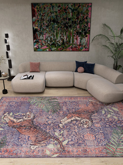 Bild 1 in Galerieansicht laden. A modern living room featuring a curved beige sofa, colorful cushions, a floor lamp, leafy plant, vibrant abstract wall art, and the Stormstripe Echo - Washable Rug - JR0046 with blue tones, tigers, and floral patterns.