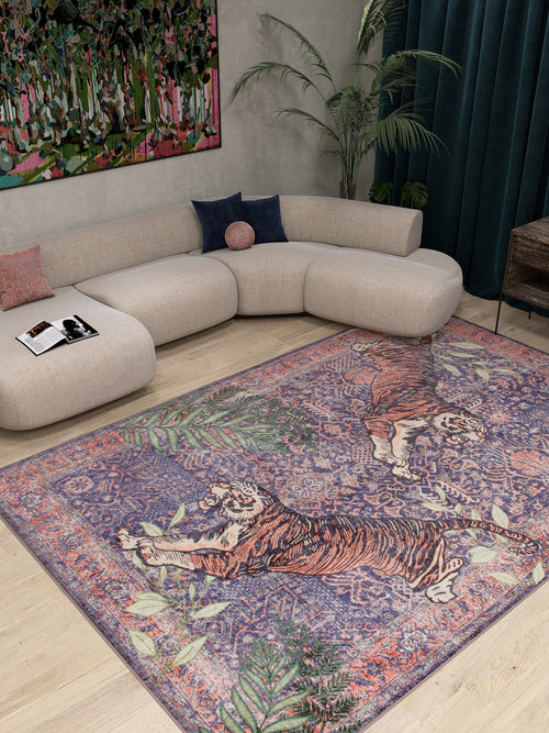 Bild 1 in Galerieansicht laden. A modern living room with a curved beige sofa, colorful cushions, a low table with a magazine, dark green curtains, tall plant, vibrant abstract painting, and the Stormstripe Echo - Washable Rug - JR0046 featuring two tigers and leafy patterns.