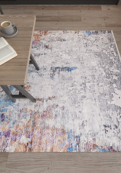 Load image 1 in gallery view. The Striking Statements - Washable Rug - JR786, featuring white, gray, blue, and copper tones, lies on a wood floor beside a light wood table with an open book and a cup of coffee.