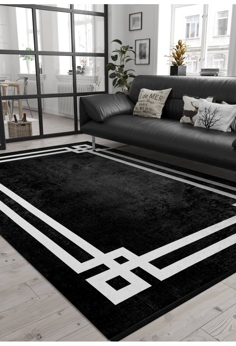 A modern living room features the Subtle Border - Washable Rug - JR597 (Outlet) US in black with a bold white border on light wood floors. Paired with a black sofa, pillows, and a potted plant, it's perfect for allergy-sensitive homes with ample natural light.