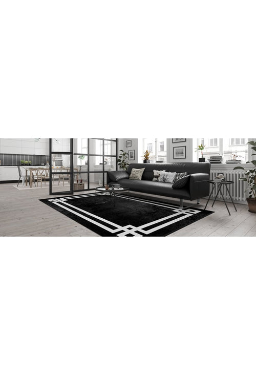Load image 1 in gallery view. A modern living room features the Subtle Border - Washable Rug - JR597 (Custom Size) in black with geometric patterns—perfect for high-traffic areas—paired with a black sofa, patterned cushions, a white coffee table, large windows, and an open kitchen-dining area.