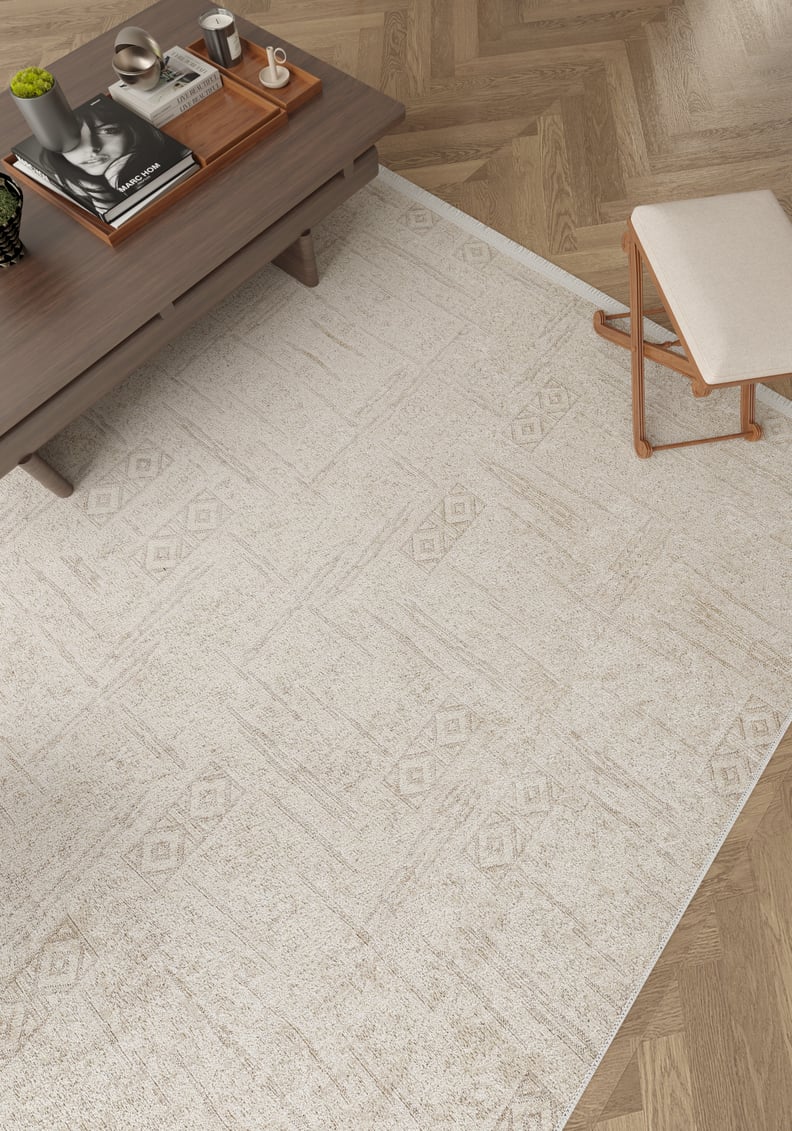 The Sunset Fade - Washable Rug - JR1878, in cream and beige, sits on a herringbone wood floor beside a wooden coffee table with books, a tray, candle, and cup—perfect for high-traffic areas—with a small upholstered stool nearby.