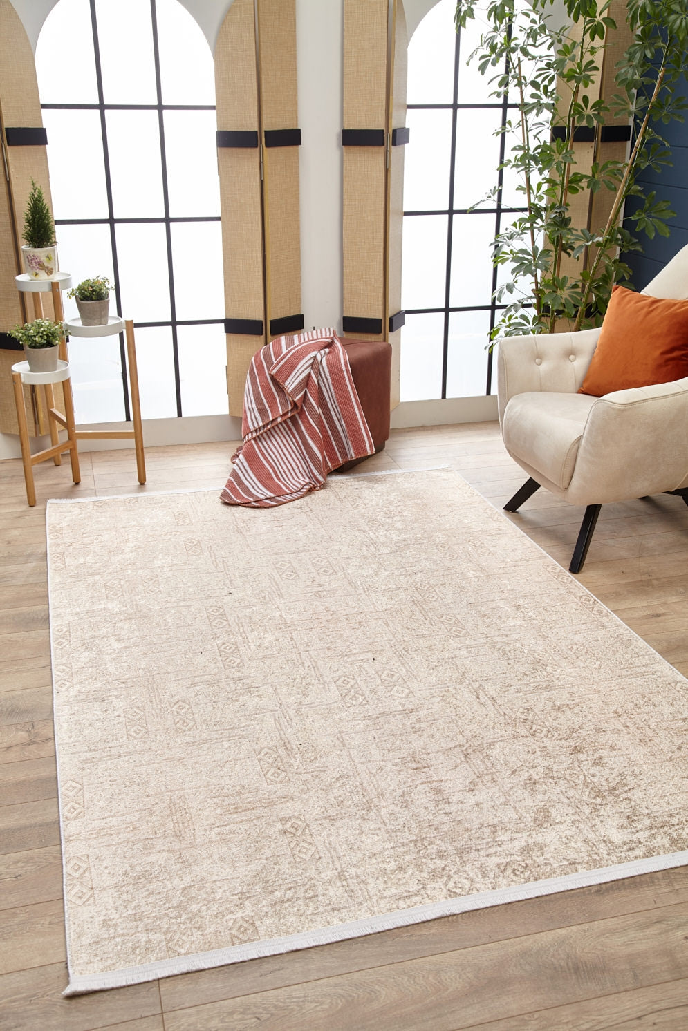 A modern living room features the Sunset Fade - Washable Rug - JR1878 (Custom Size) on wooden flooring, a cream armchair with orange pillows, potted plants on a side table, and a pouf with a red-striped blanket by tall windows.