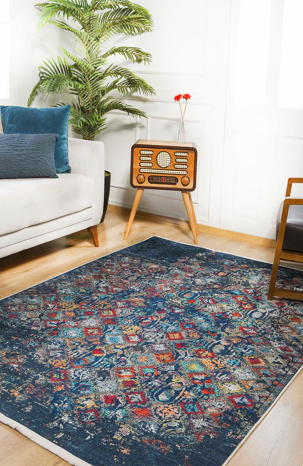 A cozy living room with the Tarnished Elegance - Washable Rug - JR1095 (Custom Size) in blue for high-traffic areas, a white sofa with blue pillows, wooden armchair, vintage radio with red flowers, potted plant, and sunlit white walls.