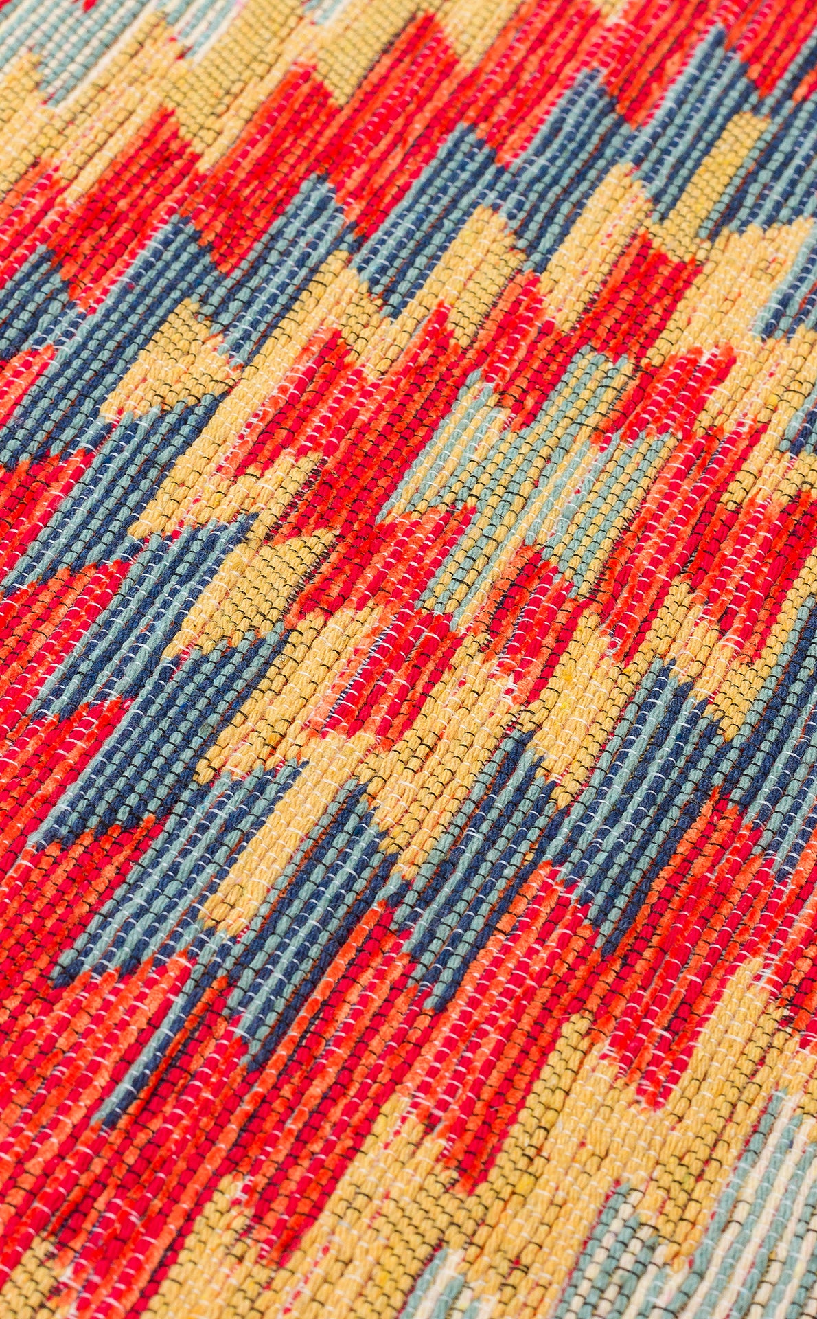 Close-up of Terra Thread - Kilim - NMD07, a flatwoven polyester fabric featuring a vibrant zigzag geometric pattern in red, yellow, and blue, inspired by tribal motifs. The detailed texture and weave are clearly visible.