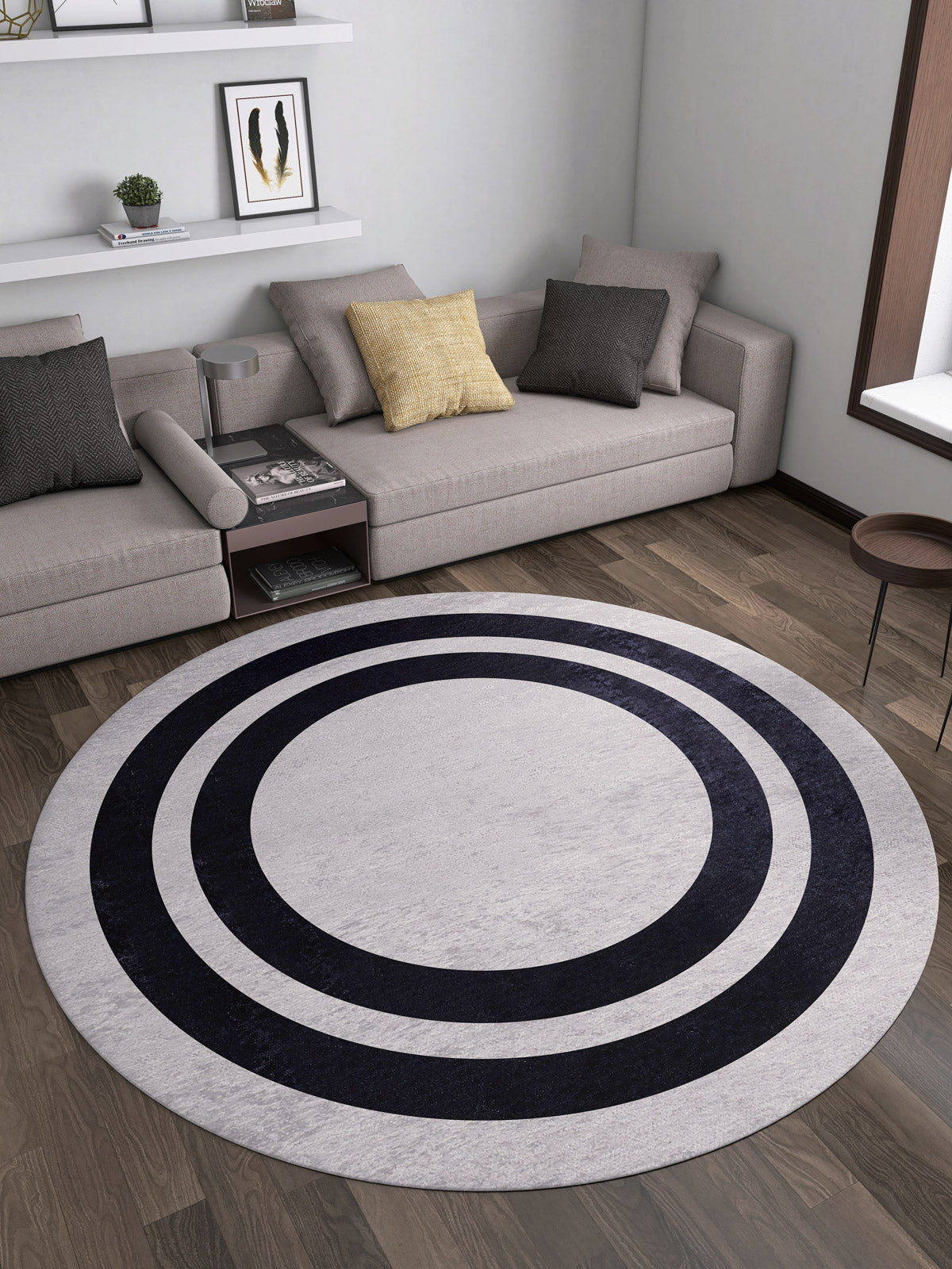 A modern living room with two beige sofas, grey and yellow cushions, the Timeless Border - Washable Rug - JR572 in gray-anthracite with black concentric circles, wooden flooring, shelves with decor, and a large window.