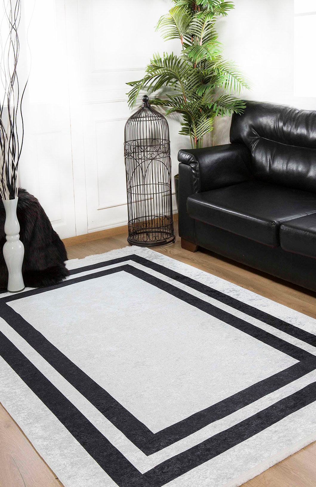 A modern living room with a black leather sofa, Timeless Border - Washable Rug - JR572 (Custom Size) in gray-anthracite with black geometric borders, a tall green potted plant, an empty black birdcage, and a white vase—perfect for high-traffic areas.