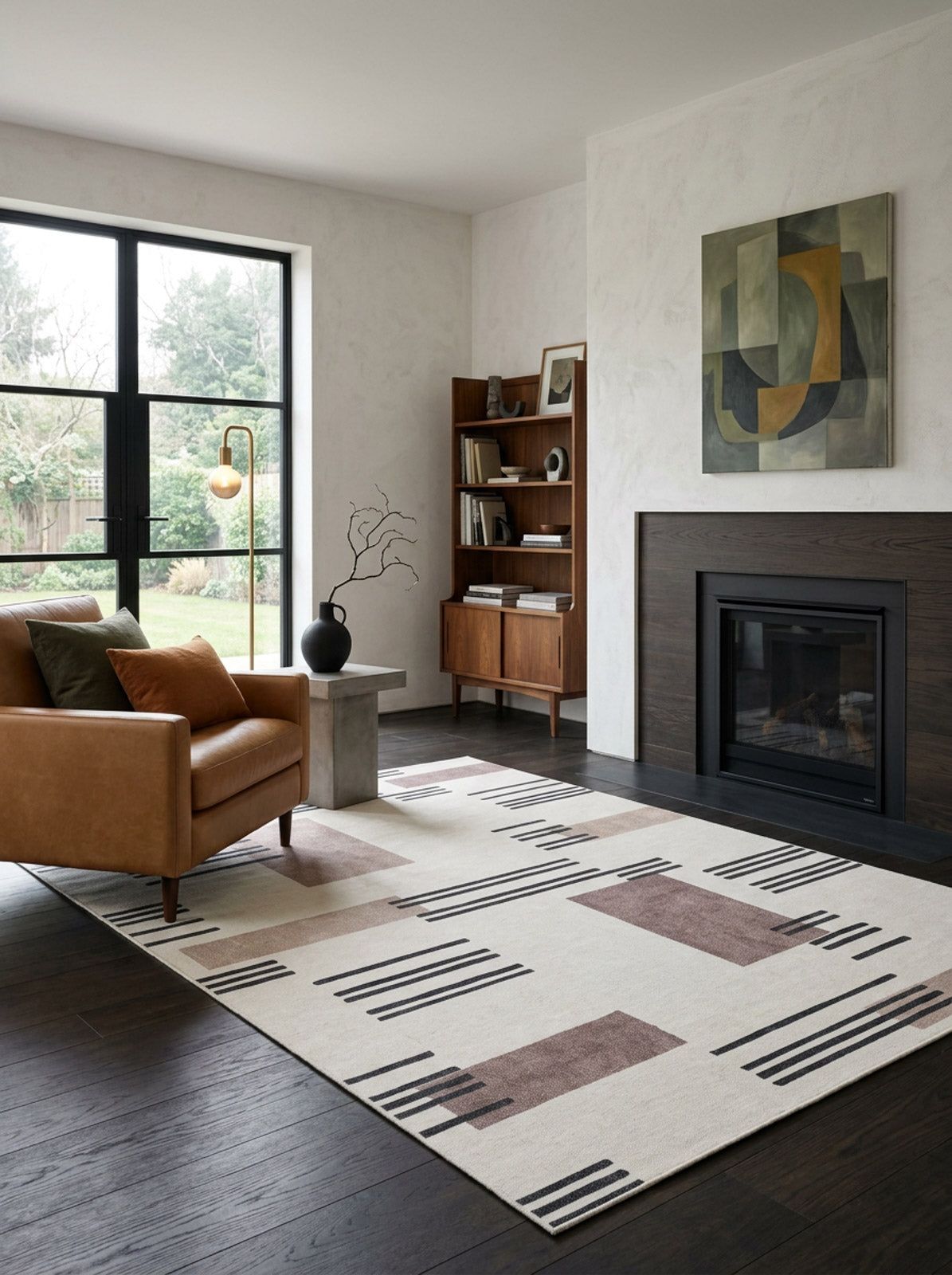 A modern living room with a tan leather sofa, the Timeless Scroll - Washable Rug - JRN45, dark wood floors, fireplace, bookshelf, and a large window overlooking greenery. An abstract painting hangs above the fireplace.