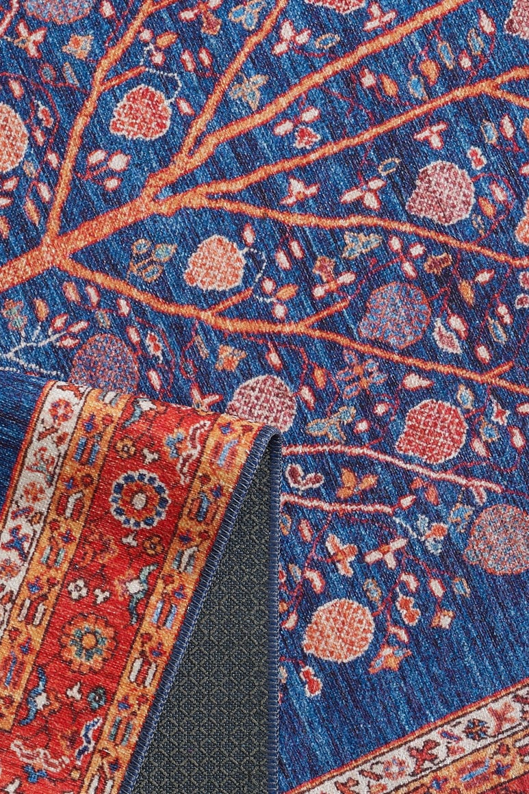 The Tree of Plenty Red Blue Washable Rug (LCC3031) features a blue background with tree branches and pomegranates, plus a red patterned border. A folded corner reveals the underside—perfect for high-traffic use.
