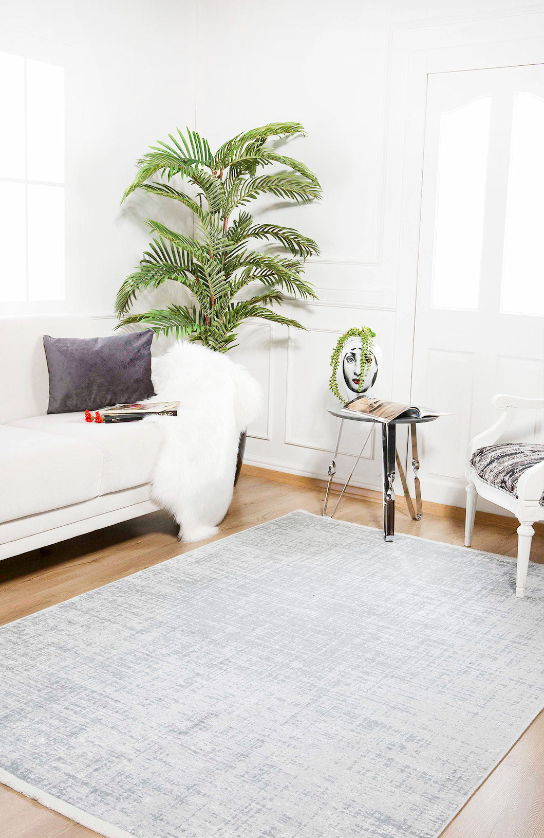 A bright, airy living room featuring a white sofa, gray pillow, fluffy throw, potted plant, modern side table with magazines and art piece, white armchair, and the Twilight Sky - Washable Rug - JR856 (Custom Size) in light gray on wooden floors.