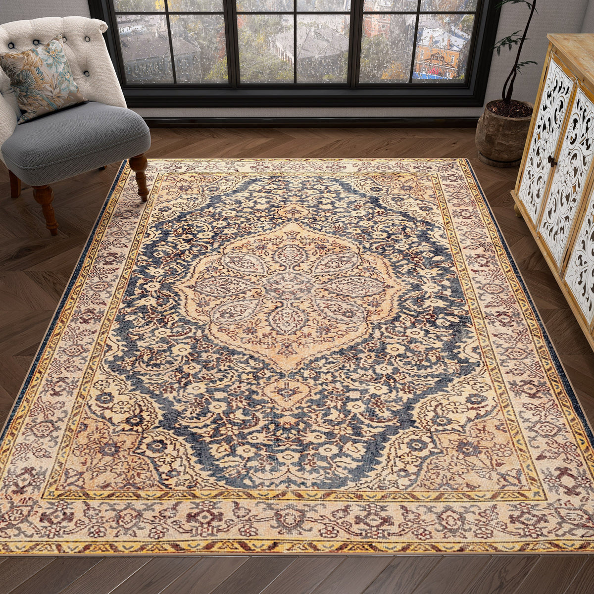 The Majestic Crest - Washable Rug - JRN1015, featuring intricate floral and medallion patterns in beige, cream, and navy blue, decorates a wooden floor in a cozy room with a cushioned chair, potted plant, and large windows.
