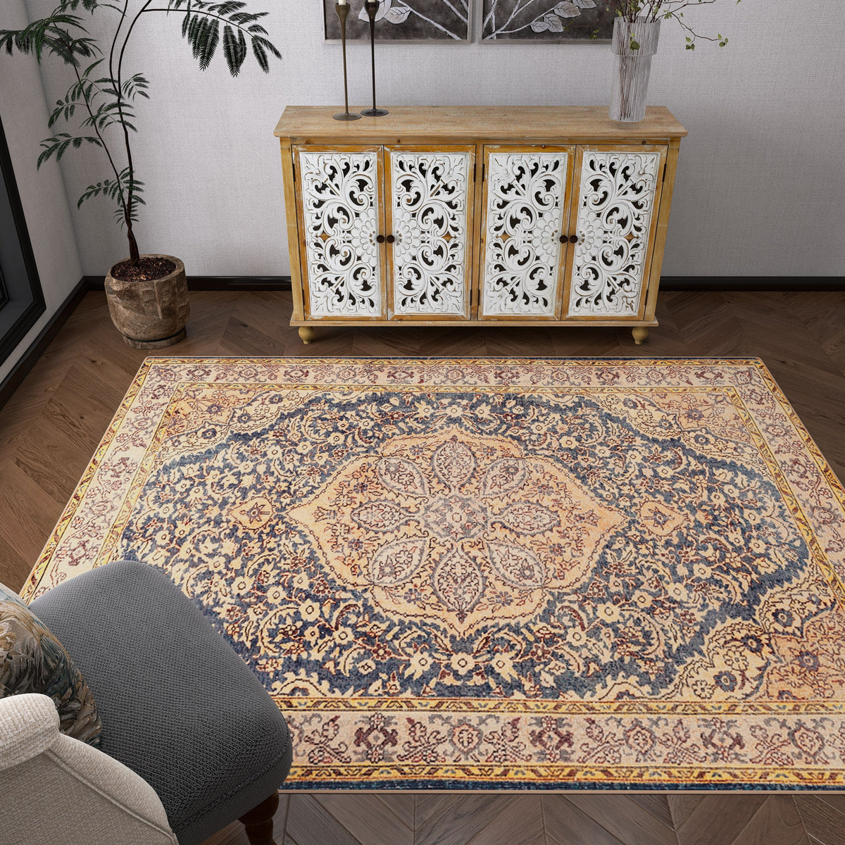 A living room features the Majestic Crest - Washable Rug - JRN1015 with floral designs in beige, blue, and gold tones, a gray armchair, a carved white and wood sideboard, potted plants, and dark herringbone wood floors.