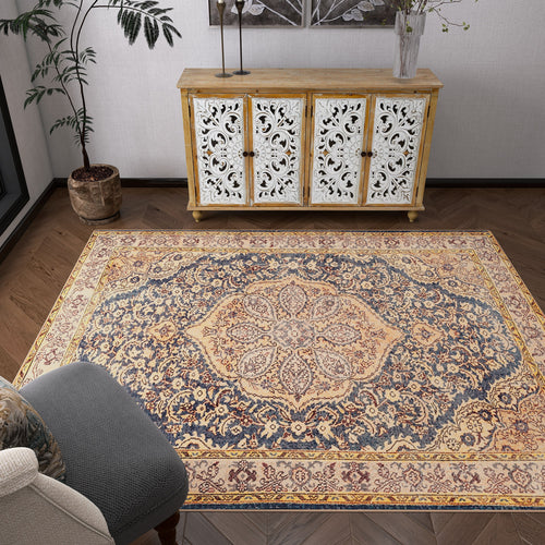 Load image 1 in gallery view. A living room features the Majestic Crest - Washable Rug - JRN1015 with floral designs in beige, blue, and gold tones, a gray armchair, a carved white and wood sideboard, potted plants, and dark herringbone wood floors.