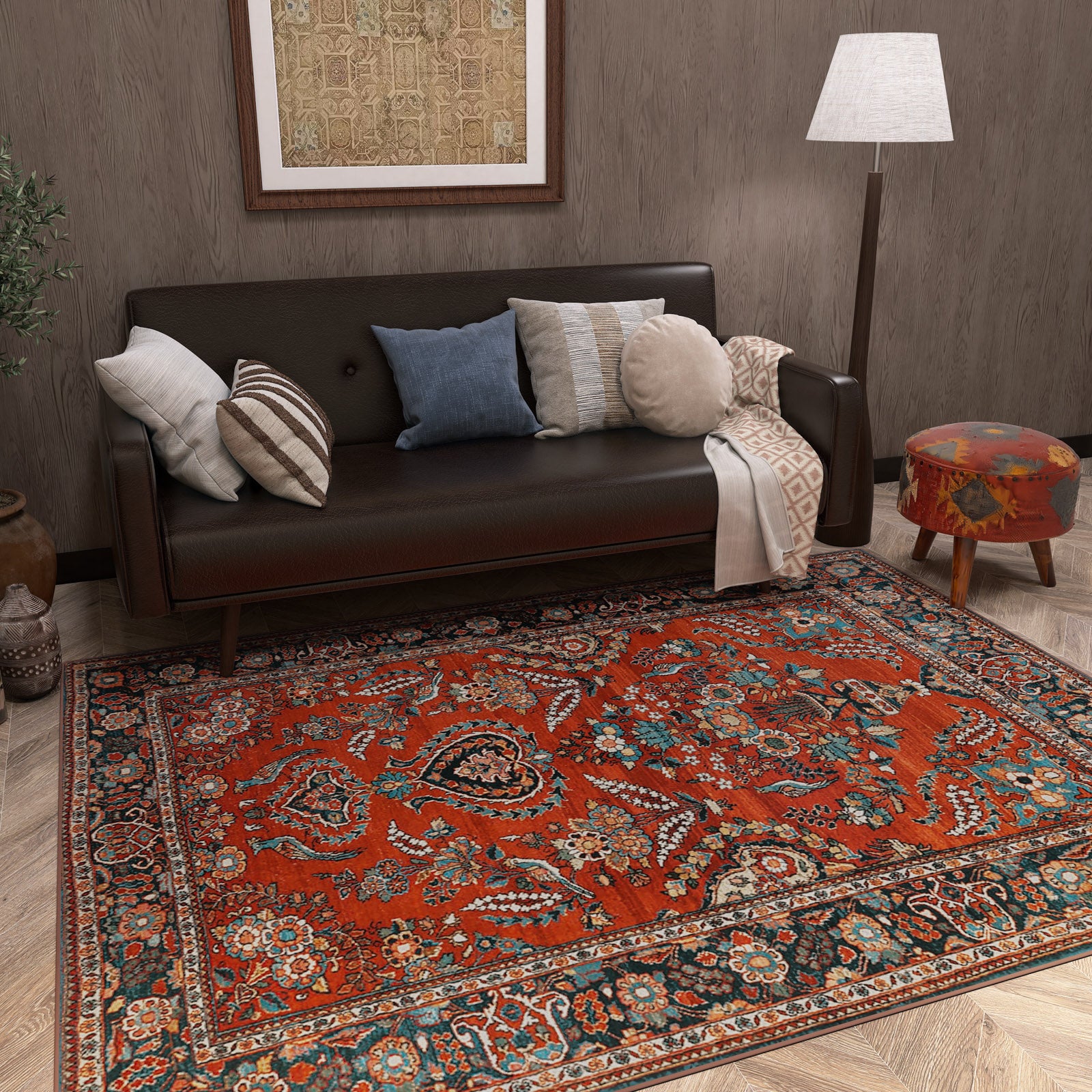 A cozy living room features a dark brown sofa with assorted pillows, a floor lamp, a colorful upholstered stool, and the Golden Scrolls - Washable Rug - JRN1005 adding vibrant patterns to the wooden floor. Artwork hangs behind the sofa.