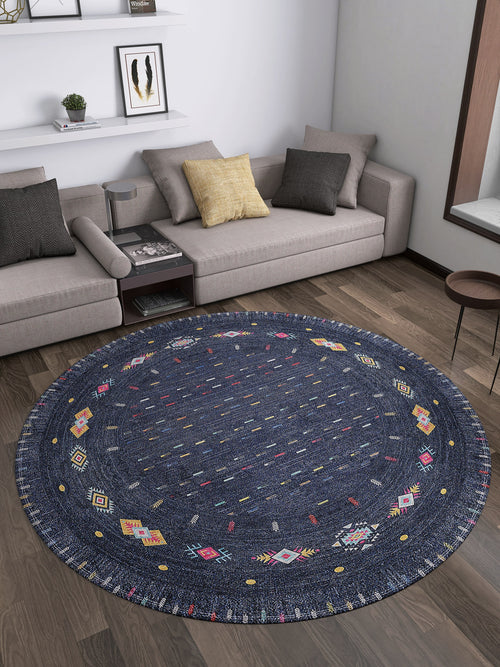 Load image 1 in gallery view. A modern living room with the Wanderer's Way - Washable Rug - JR5101 in blue, featuring colorful geometric patterns, a gray sectional sofa, assorted cushions, a round table, and wall shelves—ideal for high-traffic spaces.