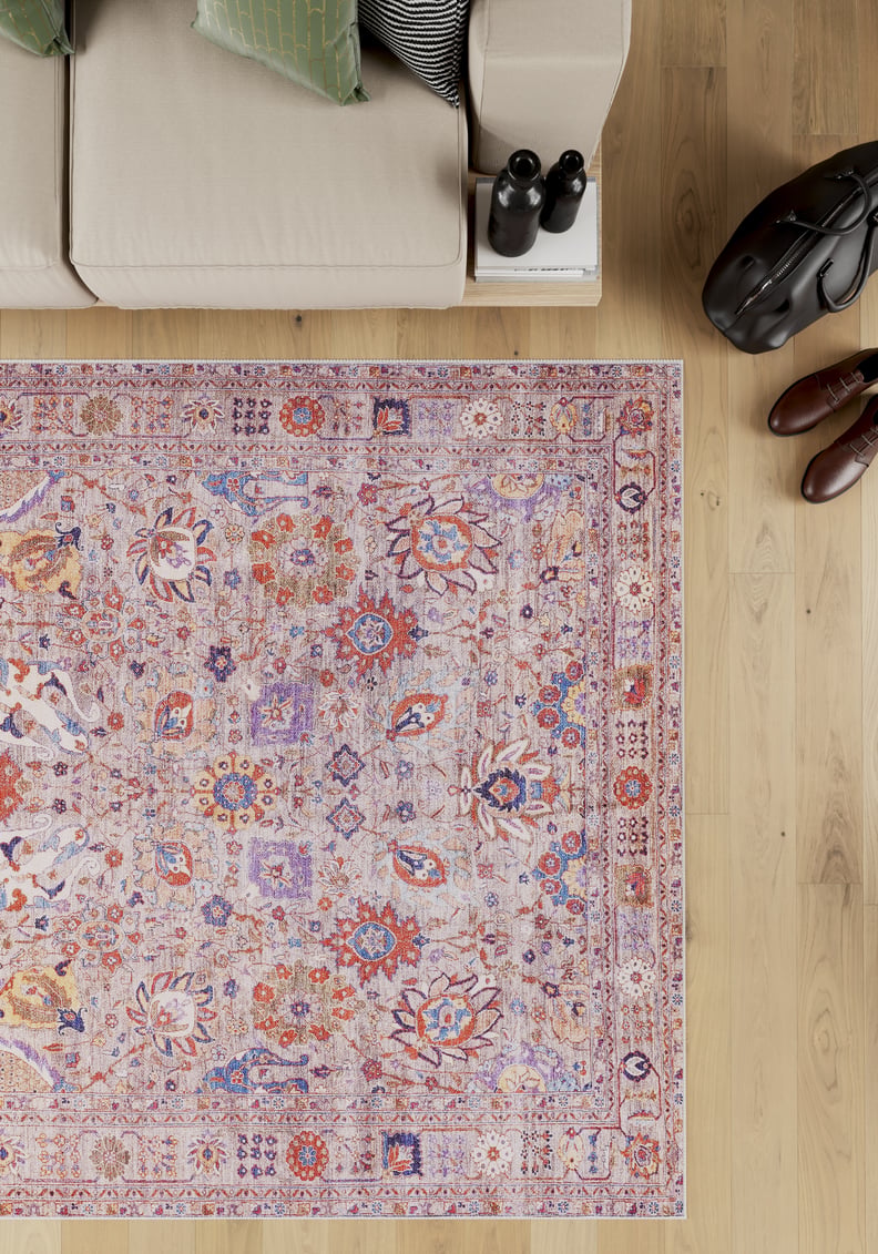 A beige sofa sits beside the Velorio Floral Washable Rug - LCC3006 (Custom Size) with pink, purple, and orange designs on a light wood floor—perfect for allergy-sensitive homes. Black binoculars, a bag, and brown shoes are nearby.