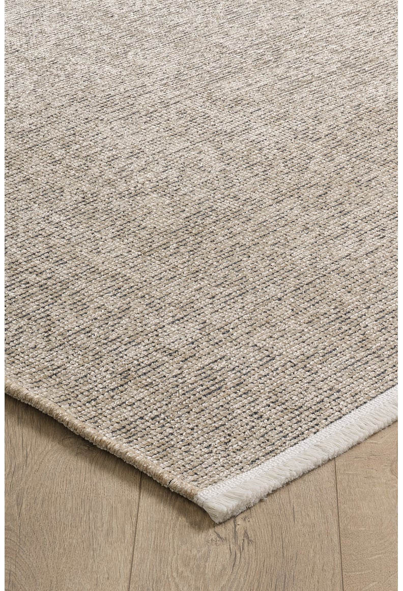 A close-up of the Velvet Touch Beige Area Rug - PST002 shows its rectangular, textured surface with a subtle woven pattern. Placed on a light wood floor, this contemporary rug features a slightly raised edge with a white border on one side.