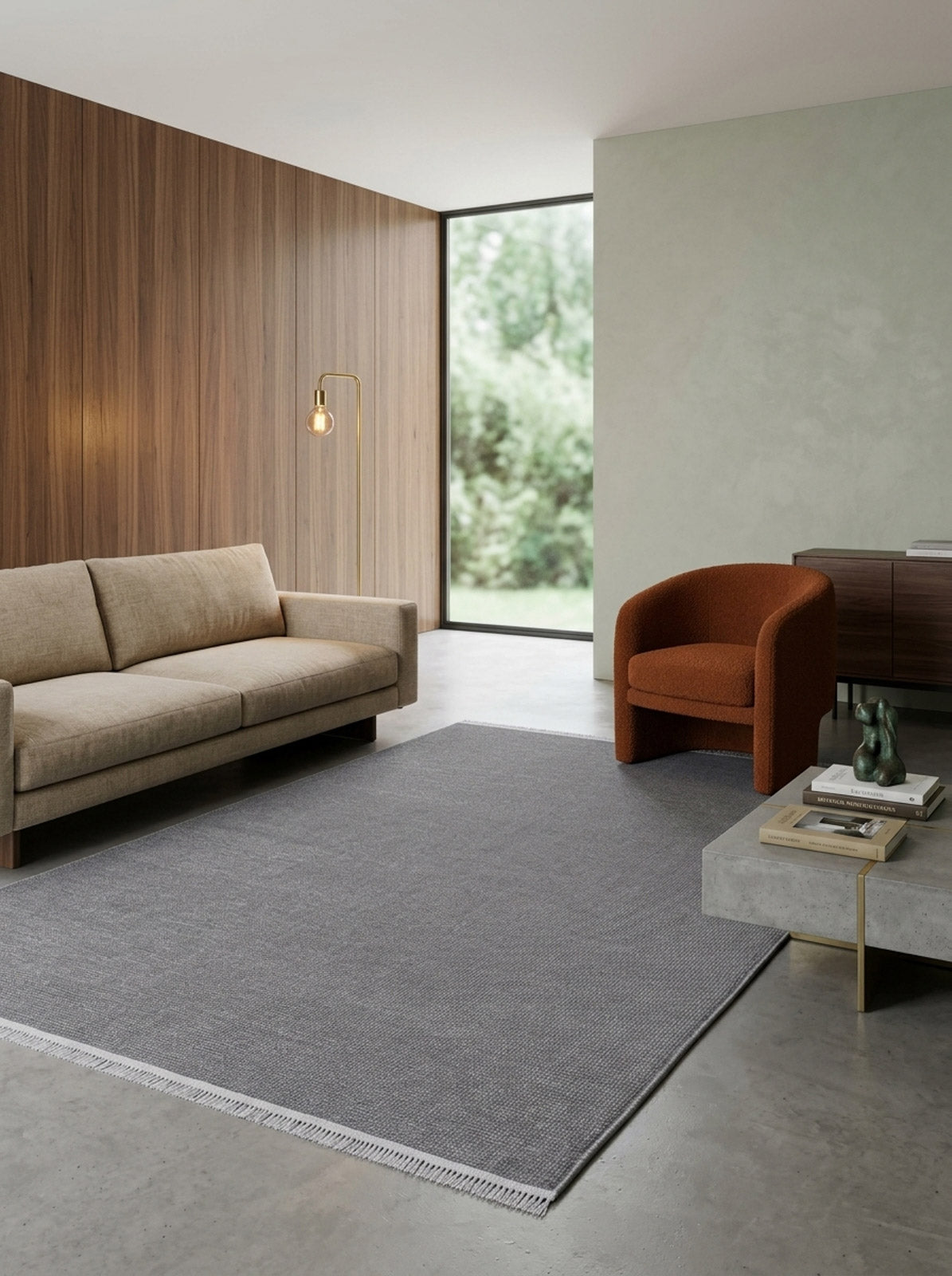 A modern living room with a beige sofa, burnt orange armchair, minimalist style, Velvet Touch Dark Gray Area Rug - PST004, concrete coffee table with books and decor, wood-paneled and green accent walls, and a large window overlooking greenery.