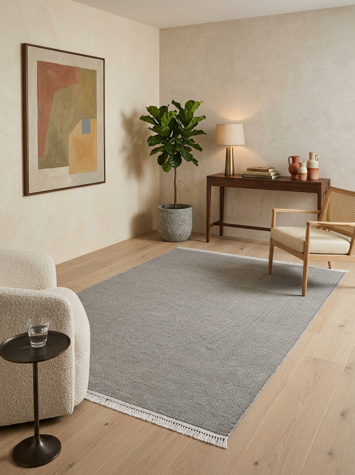 A minimalist living room with light wood floors, a cozy white chair, small side table, wooden desk, woven chair, potted plant, table lamp, abstract wall art, and the Velvet Touch Gray Area Rug - PST003 for a soft-touch feel.