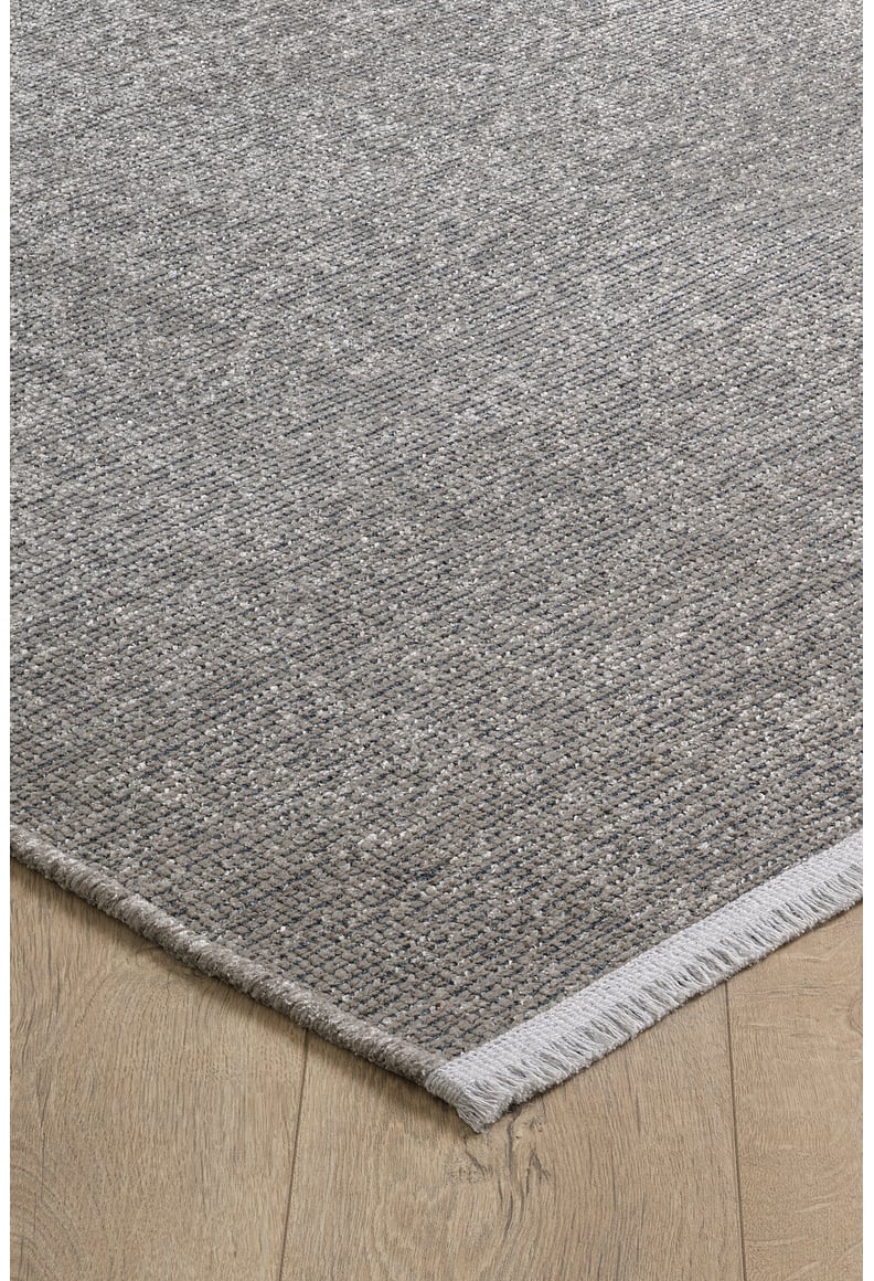 A close-up of the Velvet Touch Gray Area Rug - PST003 shows its textured, woven surface with short fringe on one edge, set on a light wood floor. Ideal for adding modern minimalist style with a speckled gray design.