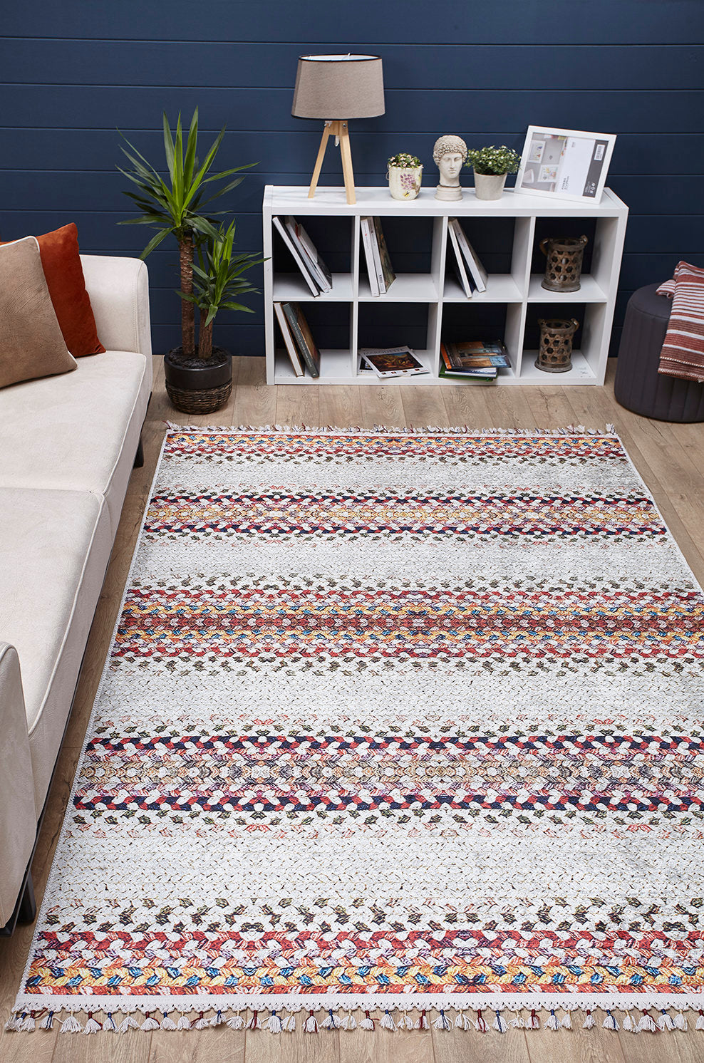 A living room featuring the Vibrant Rainbow - Machine-Washable Rug - JR5001 (Custom Size), perfect for high-traffic areas, a white sofa, potted plant, and a white shelf with books, decor, and a table lamp set against a dark blue wall.