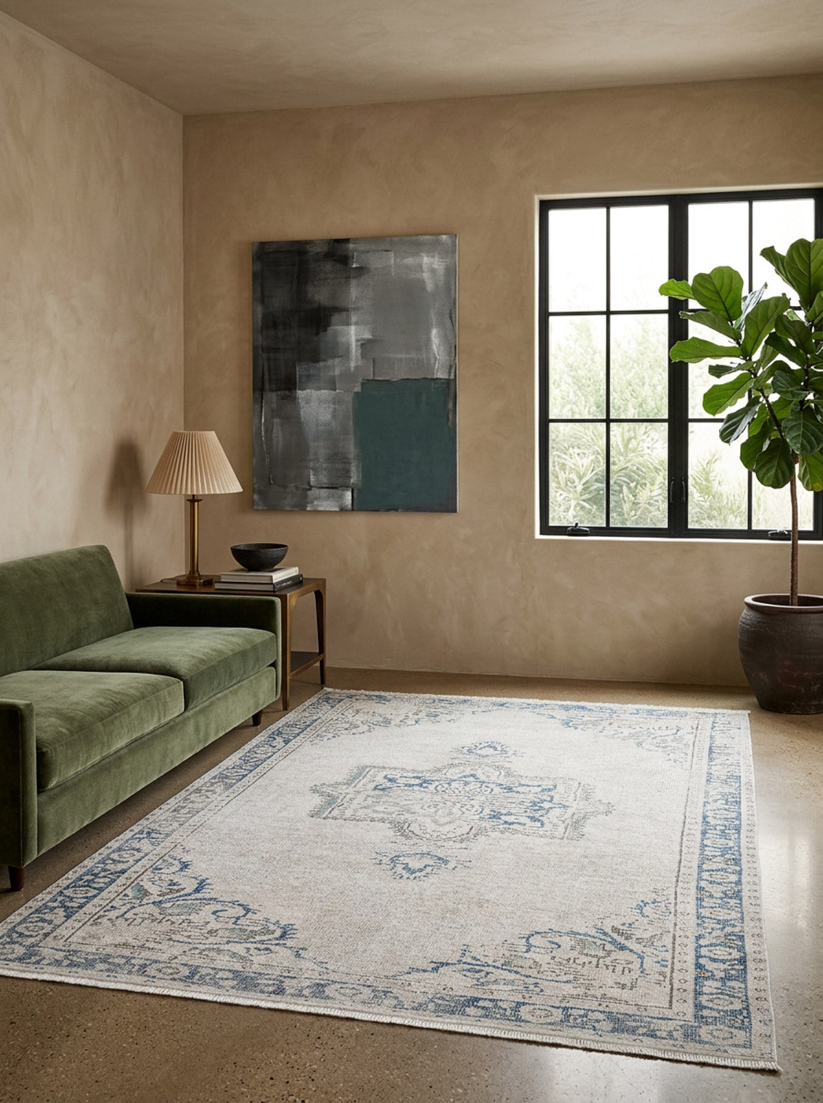 A modern living room with a green velvet sofa, the Vintage Beauty - Washable Rug - JR1688 in beige and blue, a side table holding a lamp and bowl, abstract wall art, a large potted plant, and natural light streaming through the window.