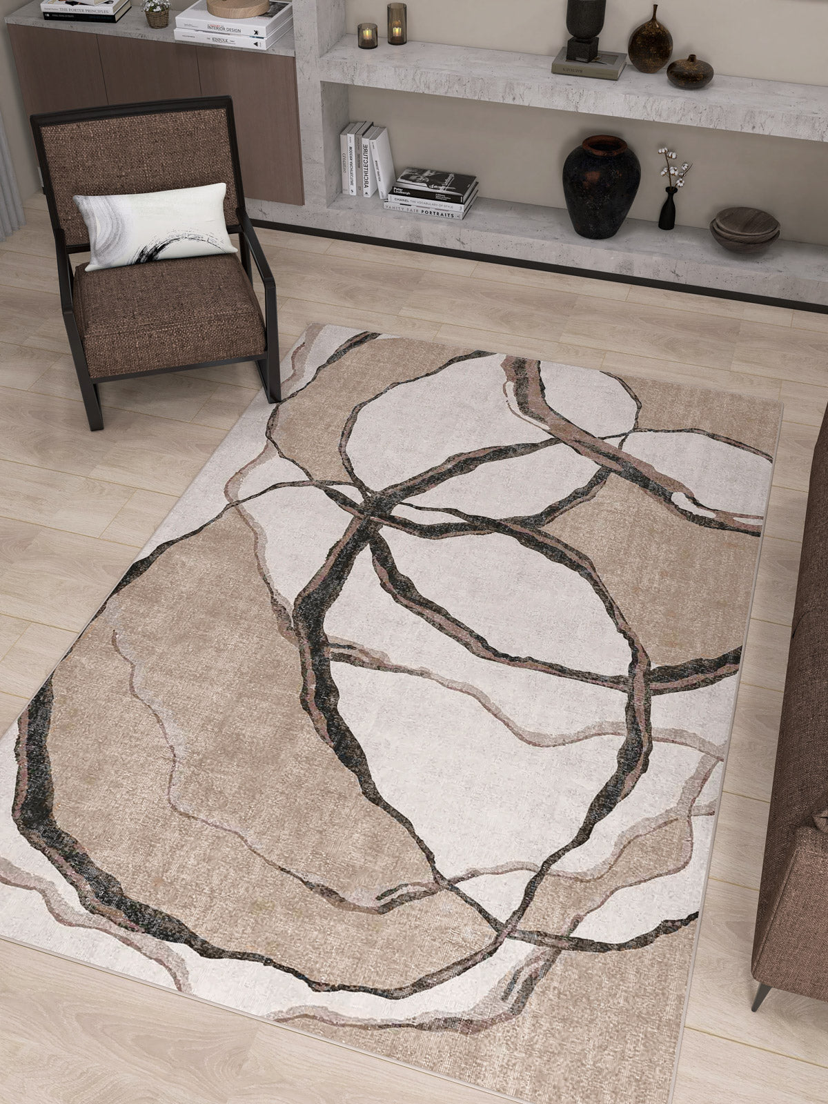 A modern living room with the Vintage Muse - Washable Rug - JRN47, a beige flat weave rug with an abstract pattern, a brown armchair with cushion, brown sofa edge, light wood floors, and shelves displaying vases and books.