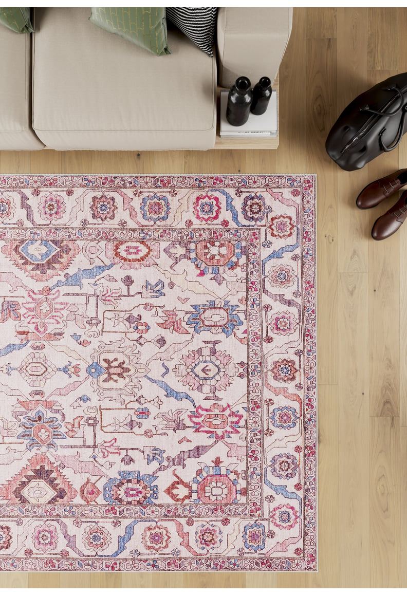 A beige sofa sits by books and a water bottle on light wood flooring near the Vivencia Authentic Washable Rug - LCC3016 (Custom Size), featuring cream, beige, pink, blue, and red patterns; a brown bag and shoes are to the right.