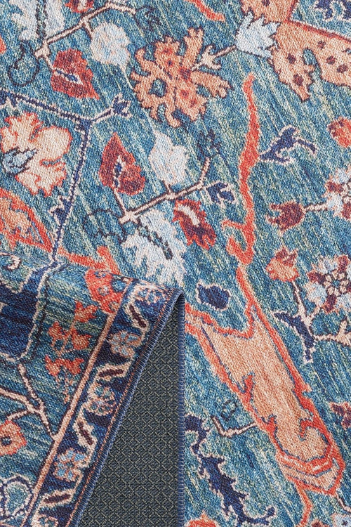 Load image 1 in gallery view. A close-up of the Whisper Authentic Blue Washable Rug - LCC3034, featuring a vibrant blue floral pattern with orange and cream accents. One corner is folded, showing the darker underside—ideal for high-traffic areas and easy to clean.