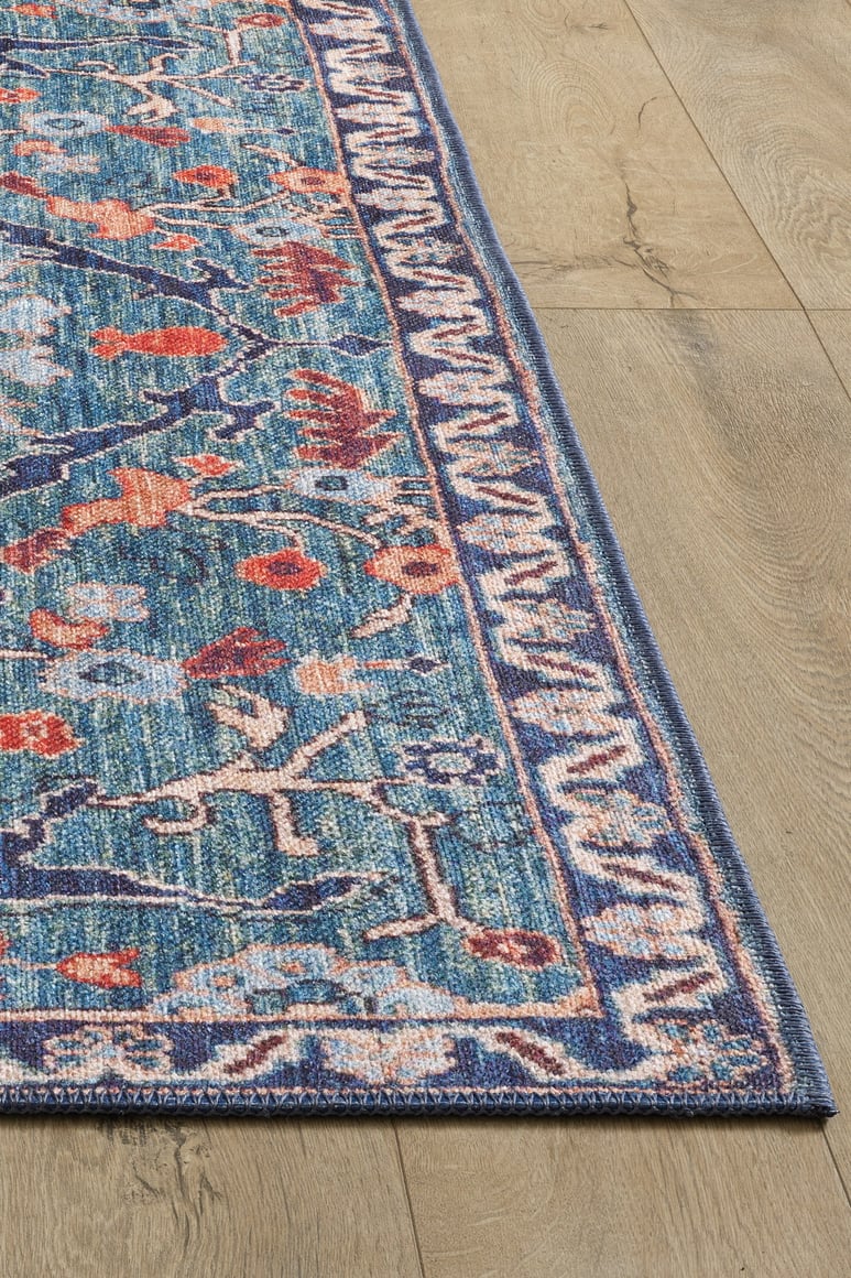 The Whisper Authentic Blue Washable Rug - LCC3034 features a blue and multicolored floral pattern with red, orange, and beige accents, plus a decorative border. It’s perfect for high-traffic areas and looks great on light wood flooring.