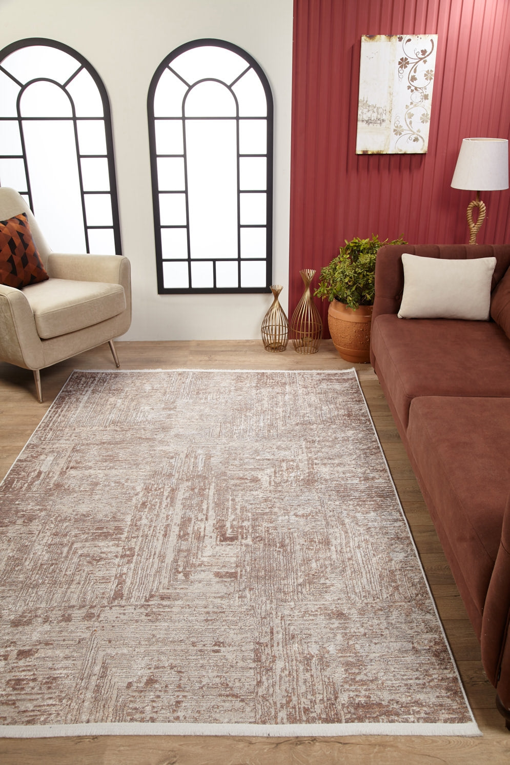 A modern living room featuring the Wildflower Fields - Washable Rug - JR1632 (Custom Size), a beige armchair, brown sofa with white pillow, potted plants, gold vases, a lamp, and arched windows on a red accent wall.
