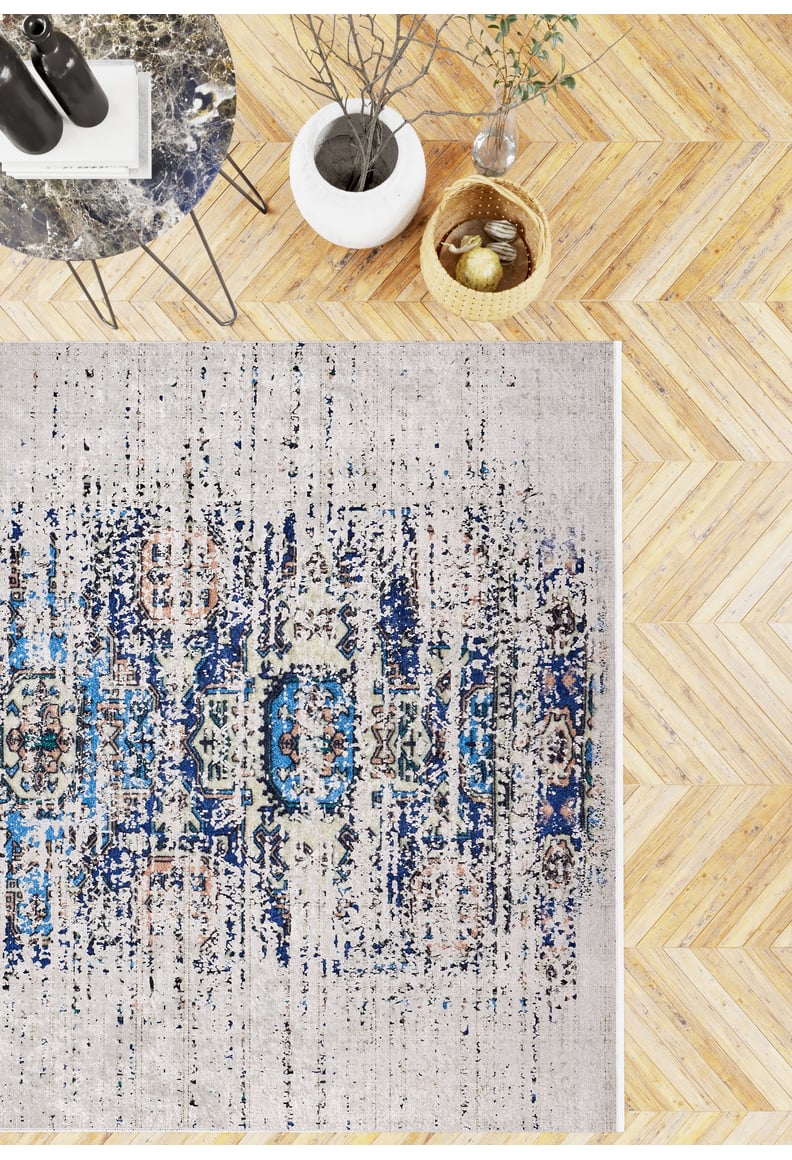 A top-down view of a light wood herringbone floor with the Worn-Out Abstract - Washable Rug - JR339 (Custom Size), a marble-topped table, white vase with branches, and a wicker basket—ideal for high-traffic areas.