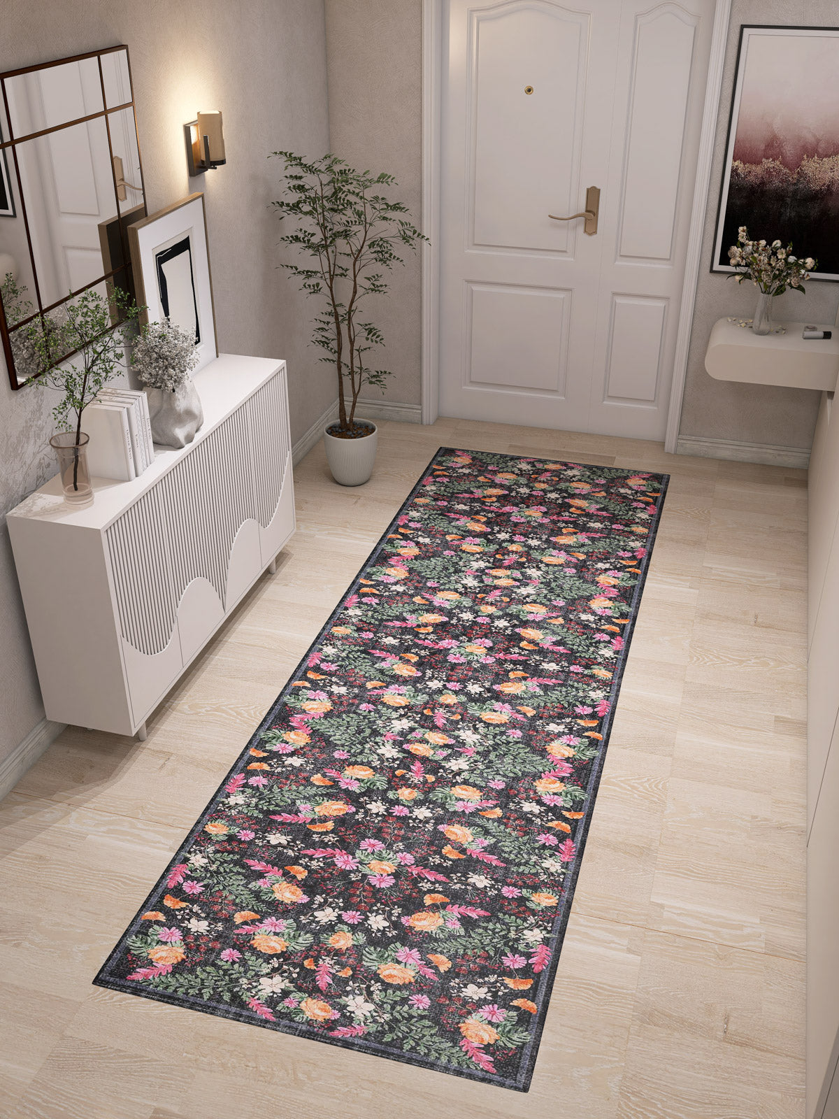 A hallway with light wood floors showcases the Woven Grace - Washable Rug - JRN28 (Custom Size), a white cabinet with decor, potted plant, and framed wall art near a white door.