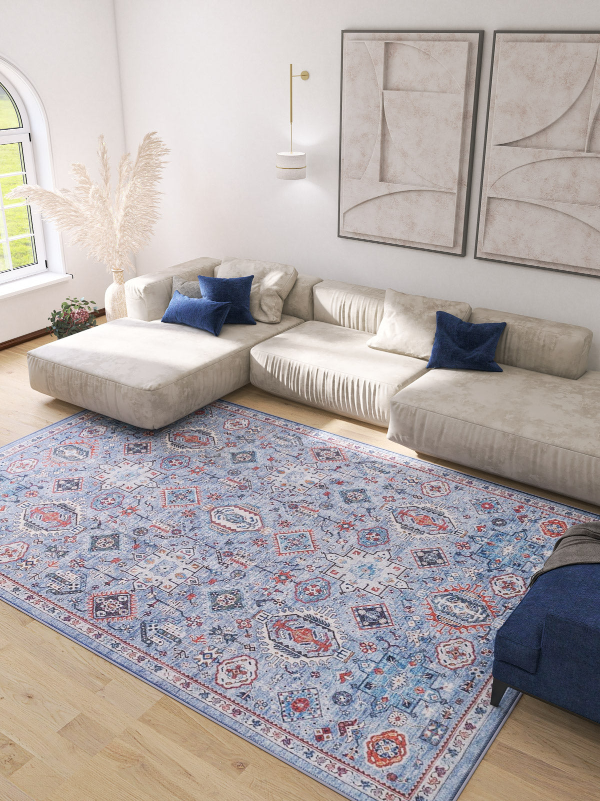 A modern living room with a beige sectional, blue accent pillows, abstract art, light wood floors, and the Xanadu Handmade-Design Blue Washable Rug – LCC3013—ideal for high-traffic areas. Pampas grass in a vase decorates the corner.