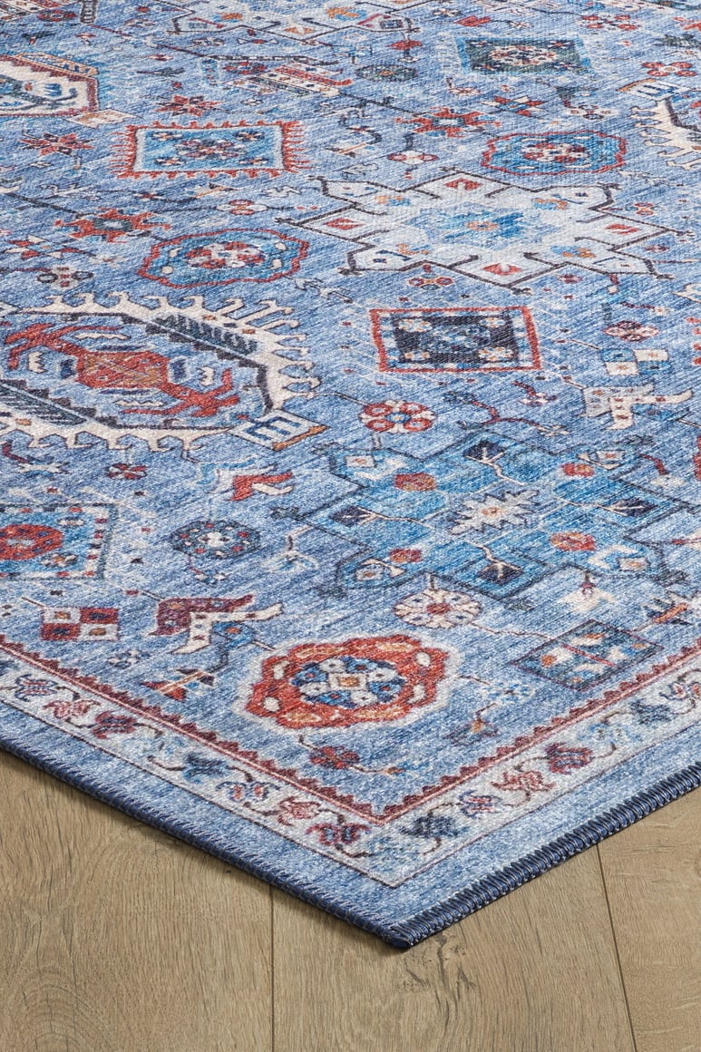 The Xanadu Handmade-Design Blue Washable Rug (LCC3013) features intricate geometric and floral patterns in red, white, and navy on a blue background, shown on a light wood floor with a decorative border—perfect for high-traffic areas.