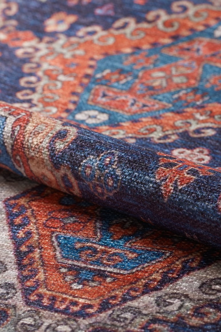 A close-up shows the Zephyro Kilim-Design Washable Rug - LCC3008, folded to display its intricate geometric pattern in blue, orange, red, and cream. The visible weave texture highlights its durability—perfect for high-traffic spaces.
