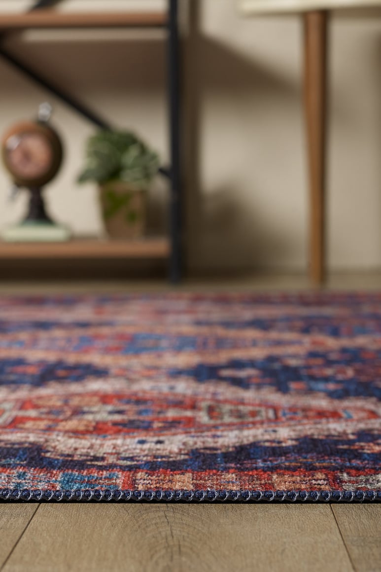 A close-up of the Zephyro Kilim-Design Washable Rug - LCC3008, featuring red, blue, and beige hues on a wooden floor. In the blurred background are a plant, decor piece, and part of a wood-legged table—perfect for high-traffic spaces.