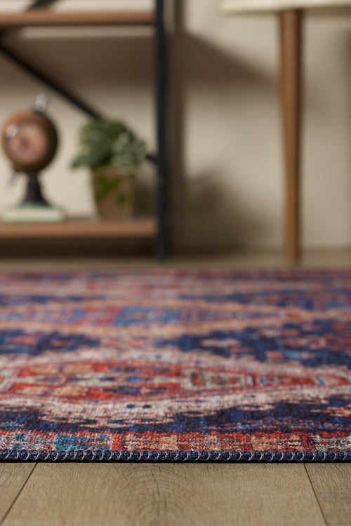 Load image 1 in gallery view. A close-up of the Zephyro Kilim-Design Washable Rug - LCC3008 (Custom Size), featuring vibrant red, blue, and cream tones on a wooden floor. Great for high-traffic areas, with blurred furniture and a potted plant in the background.