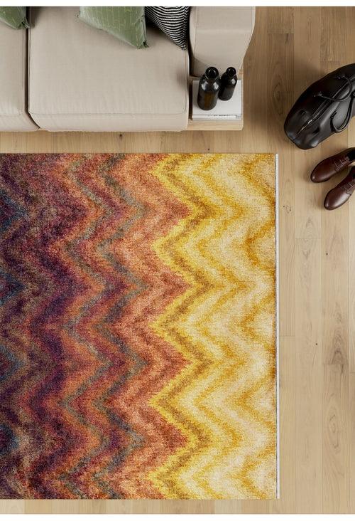 Bild 1 in Galerieansicht laden. The Zigzag Runway - Washable Rug - JR388, featuring vibrant yellow, orange, brown, and purple zigzag patterns, lies on a wooden floor beside a beige sofa. Brown shoes, a black bag, and books add to the cozy atmosphere—perfect for high-traffic areas.
