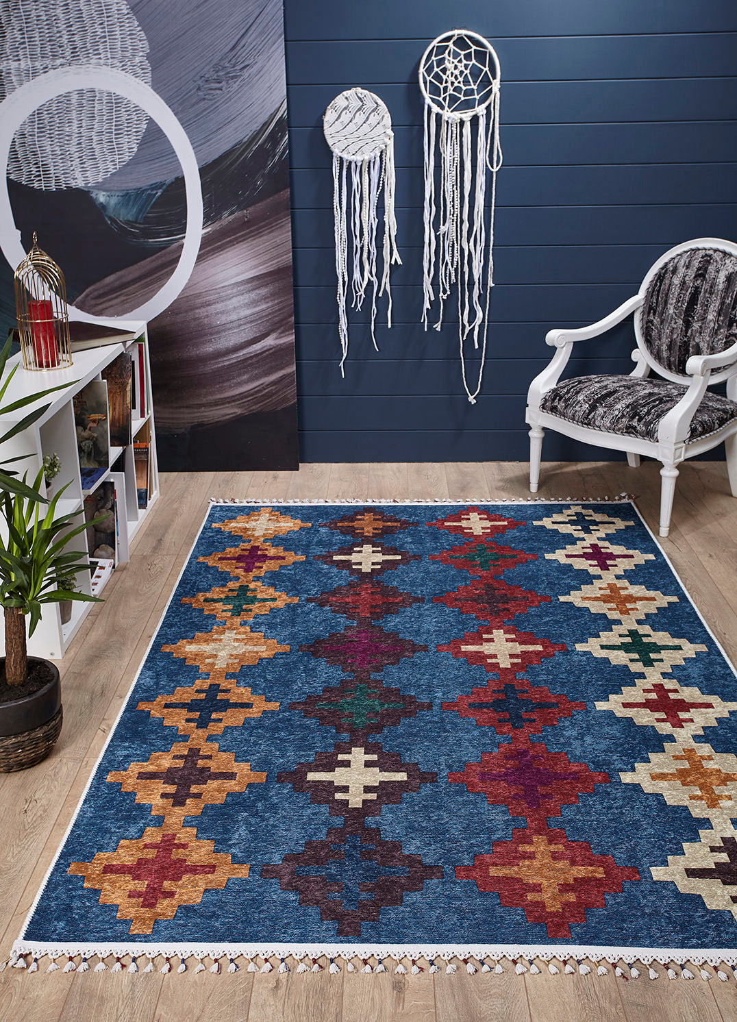 A room with a Nomad's Mirage - Washable Rug - JR5032 (Custom Size) on wooden flooring, a white armchair with a patterned cushion, wall art and dreamcatchers on a dark blue wall, plant, and white bookshelf—ideal for high-traffic areas.