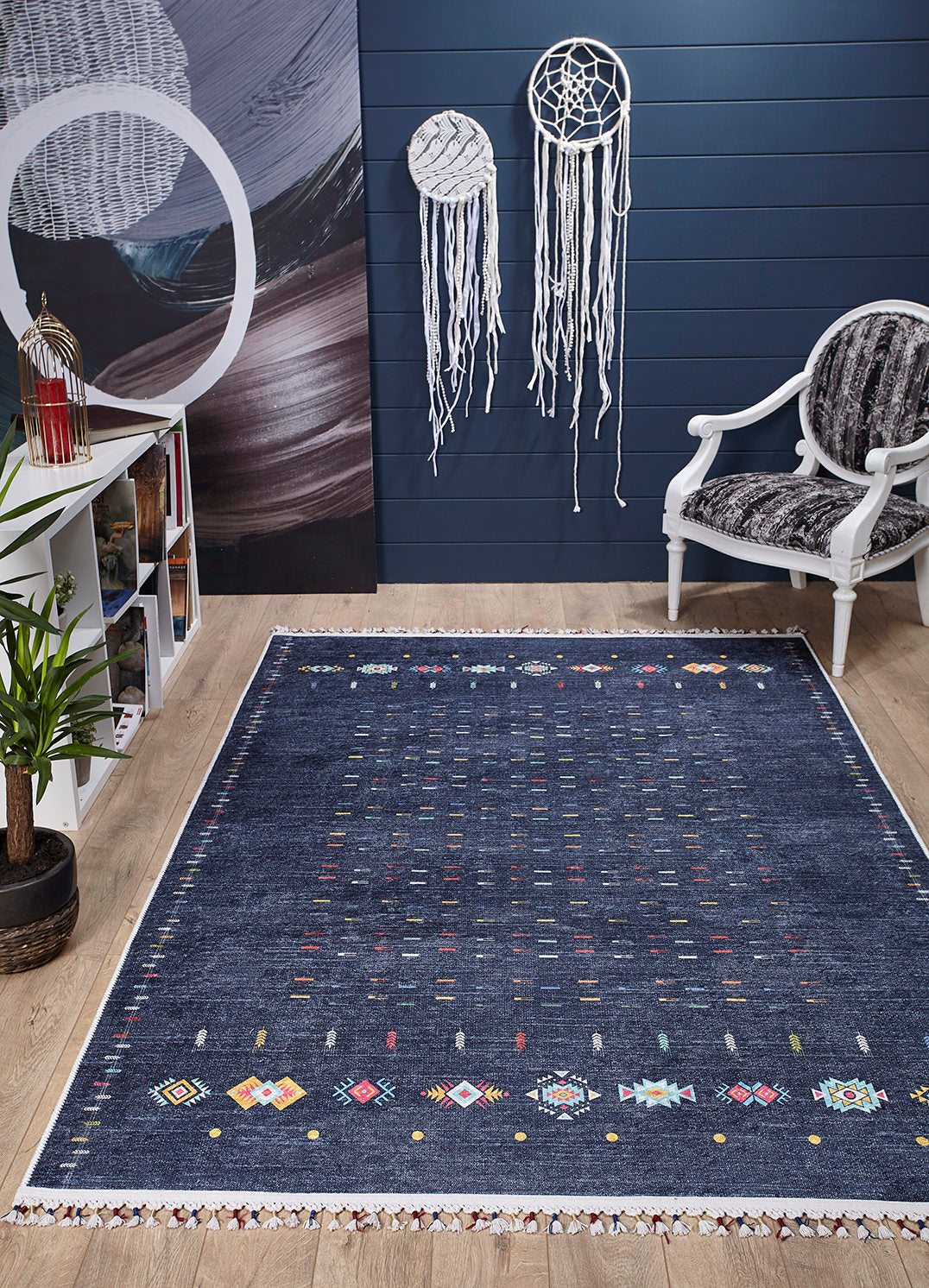 A modern room with the Wanderer's Way - Washable Rug - JR5101 (Custom Size) in blue, a white armchair with dark cushion, dreamcatchers on a navy wall, white shelf with books and decor, potted plant, and light wood floor—ideal for high-traffic spaces.