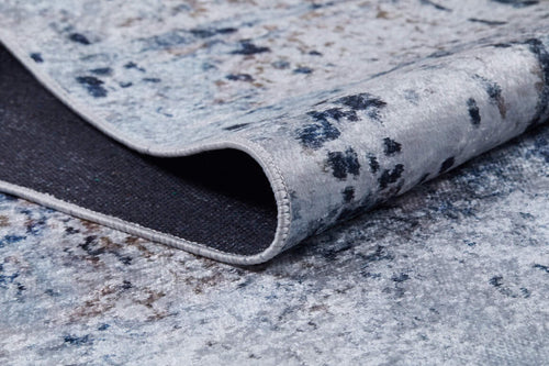 Load image 1 in gallery view. A close-up of the Abstract Atmosphere - Washable Rug - JR834 in light grey and blue, featuring an abstract pattern with a slightly lifted edge revealing the dark underside and stitched border.