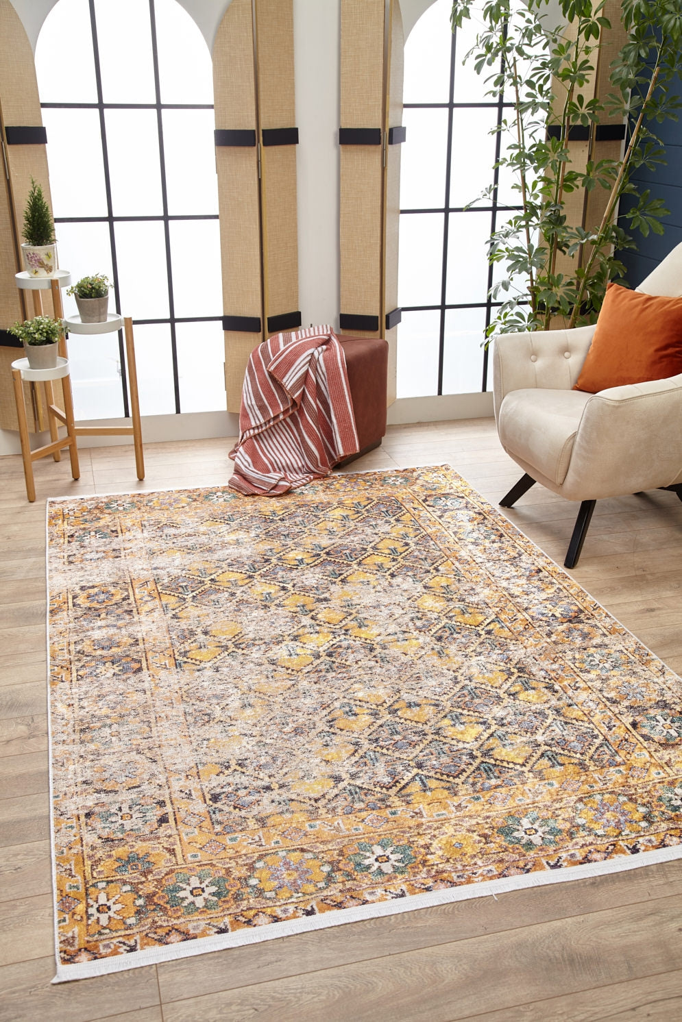 A cozy living room features the Aged Splendor - Washable Rug - JR1906 in yellow-gold, a cream armchair with an orange cushion, a brown ottoman with a striped blanket, potted plants, and large arched windows flooding the space with natural light.
